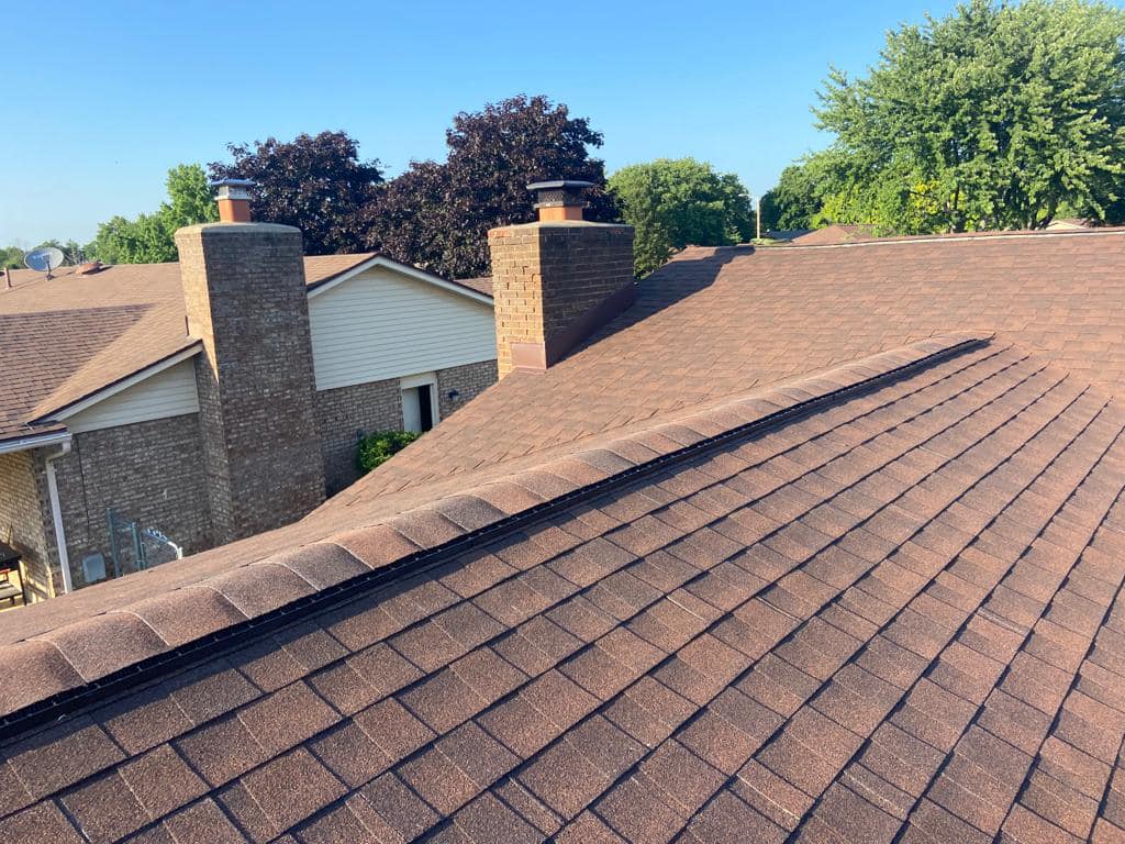 Brown asphalt shingle roof with ridge vent in residential neighborhood with brick chimneys and mature trees
