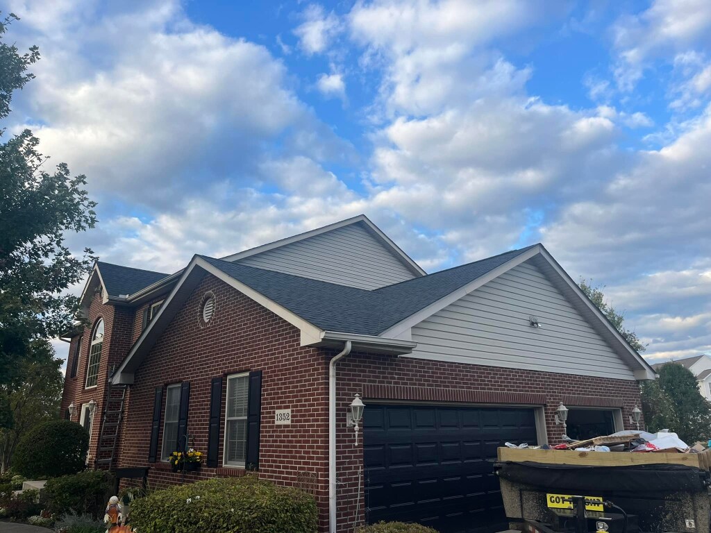 Red brick house with gray siding and dark roof under blue cloudy sky with dumpster in front yard