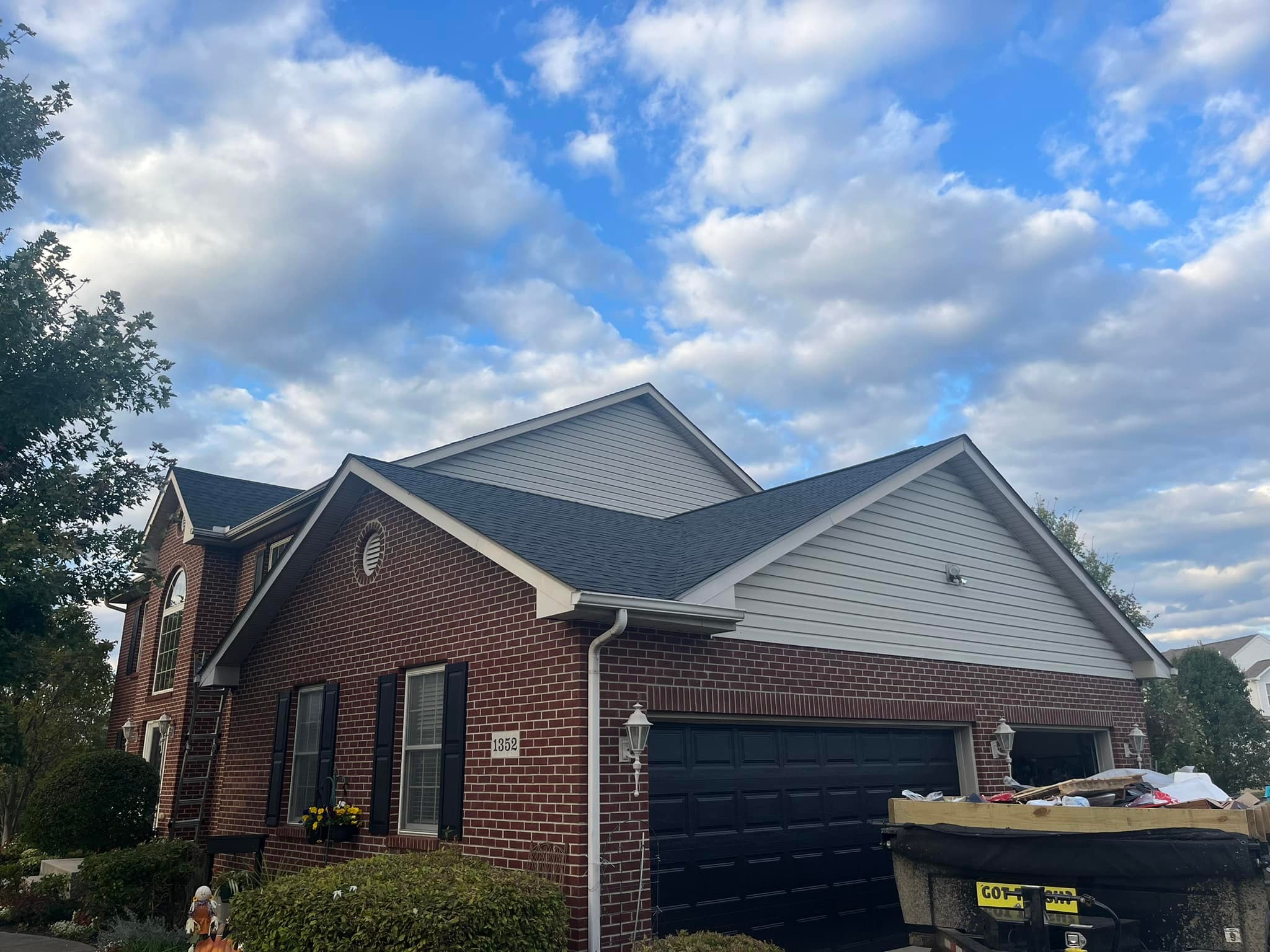 Red brick house with gray siding and dark roof under blue cloudy sky with dumpster in front yard