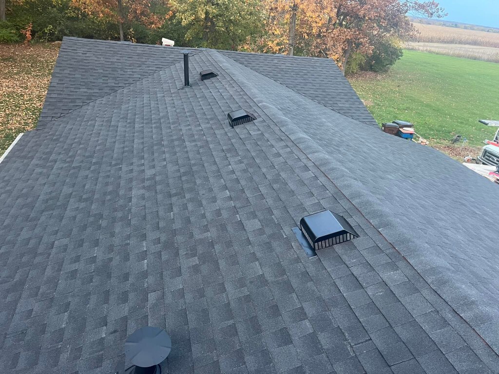 Gray asphalt shingle roof with multiple black roof vents and chimney pipe, surrounded by autumn trees and lawn