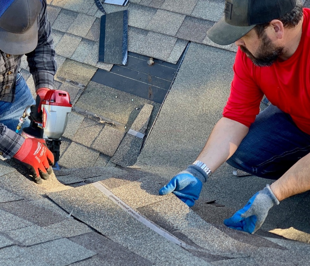 Two roofers repairing asphalt shingles on residential roof using power drill and hand tools for maintenance work