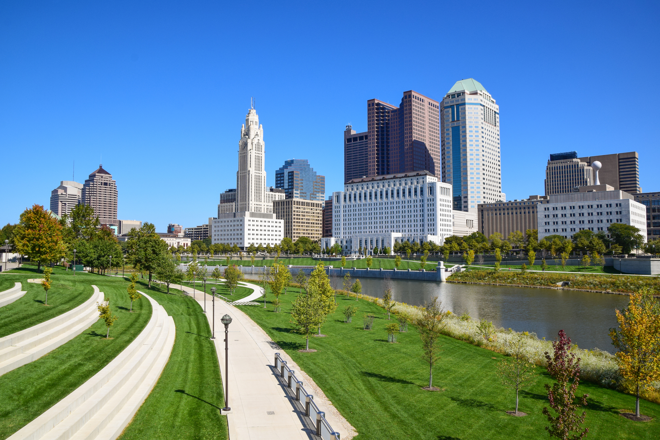Columbus Ohio downtown skyline with modern skyscrapers, green park walkways, and Scioto River under clear blue sky