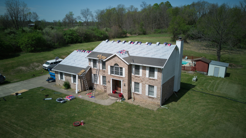 Aerial view of two-story brick house with new roof installation, workers and materials visible on rooftop