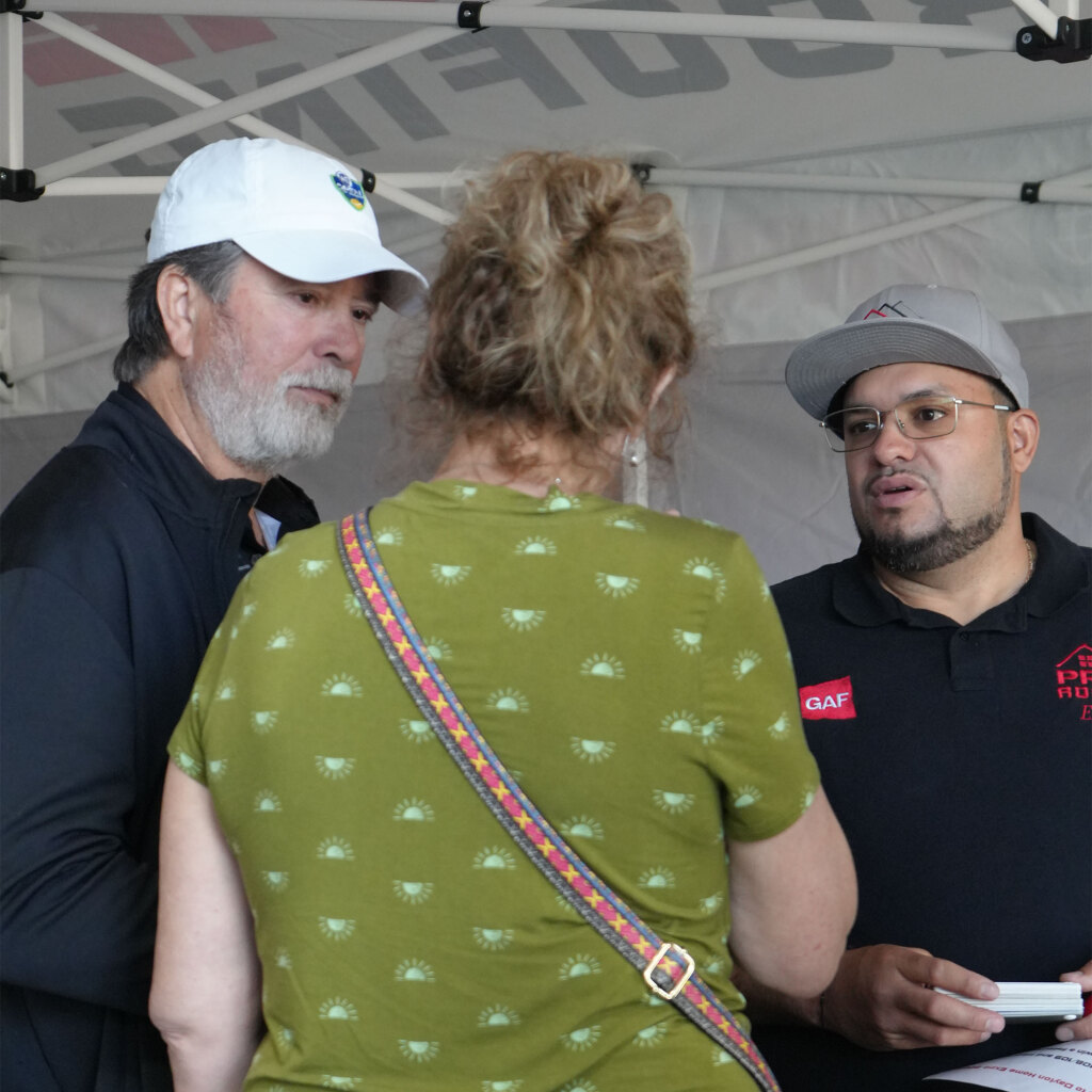 Three people having conversation under white tent, man in white baseball cap, woman in green floral top, man in gray cap