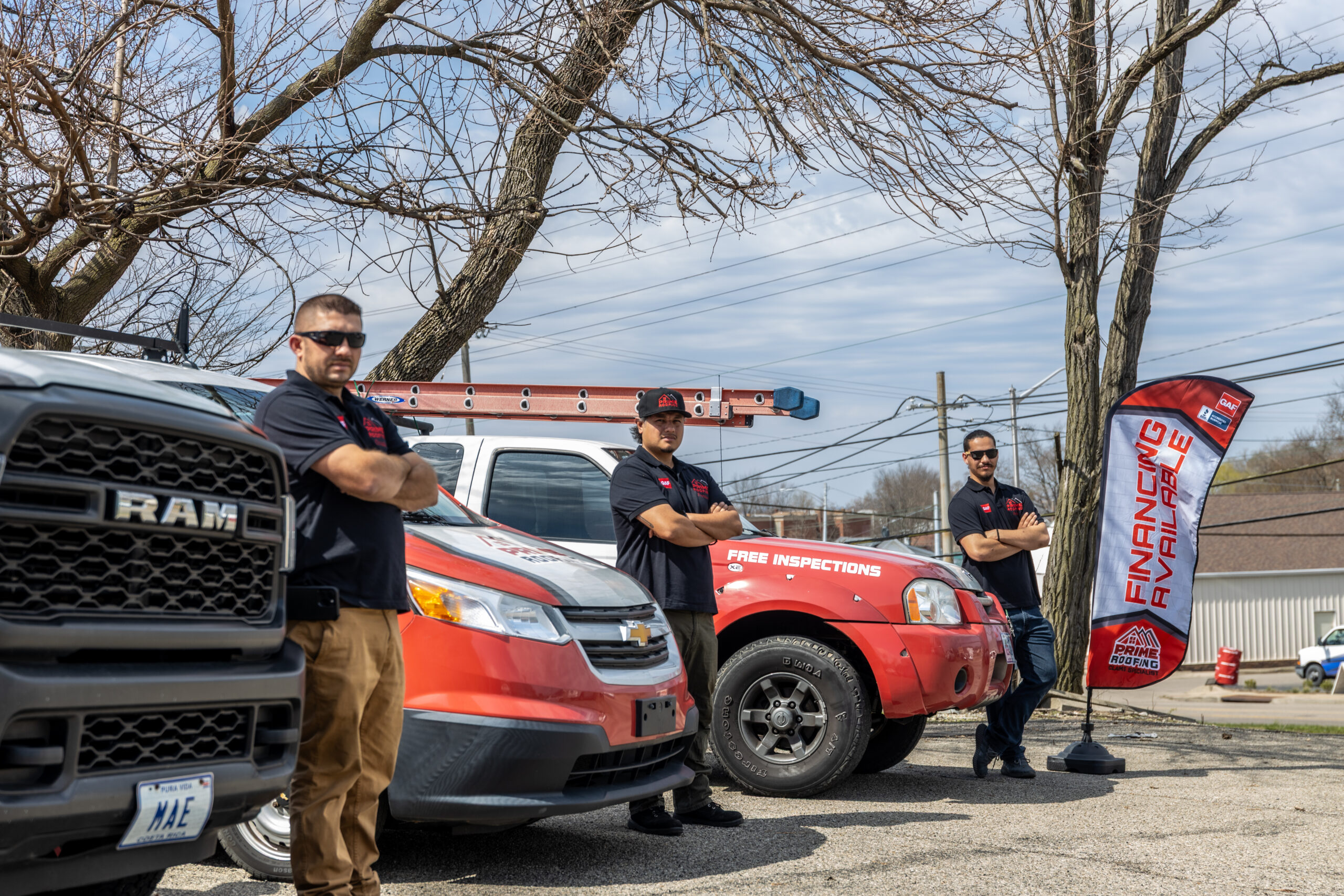 Three roofing contractors in matching uniforms standing with arms crossed next to work trucks and ladder equipment