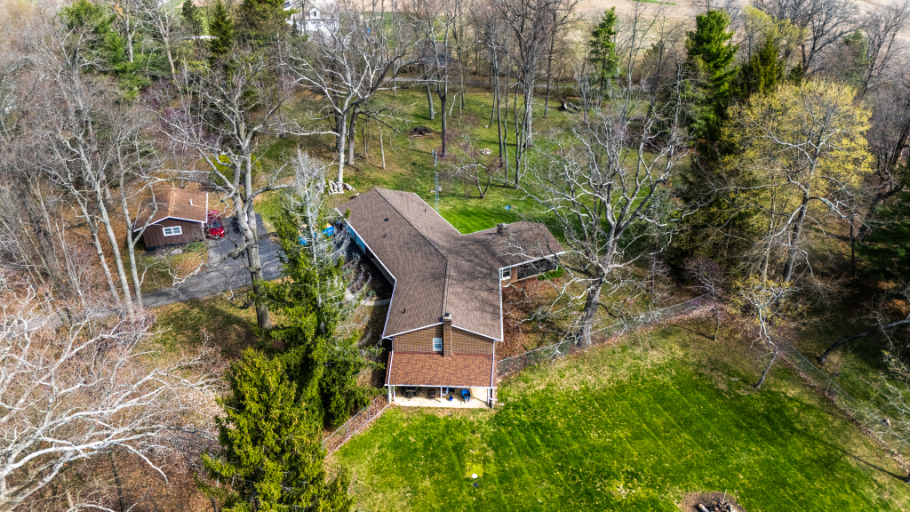 Aerial view of residential home with brown roof surrounded by green lawn and mixed deciduous evergreen trees