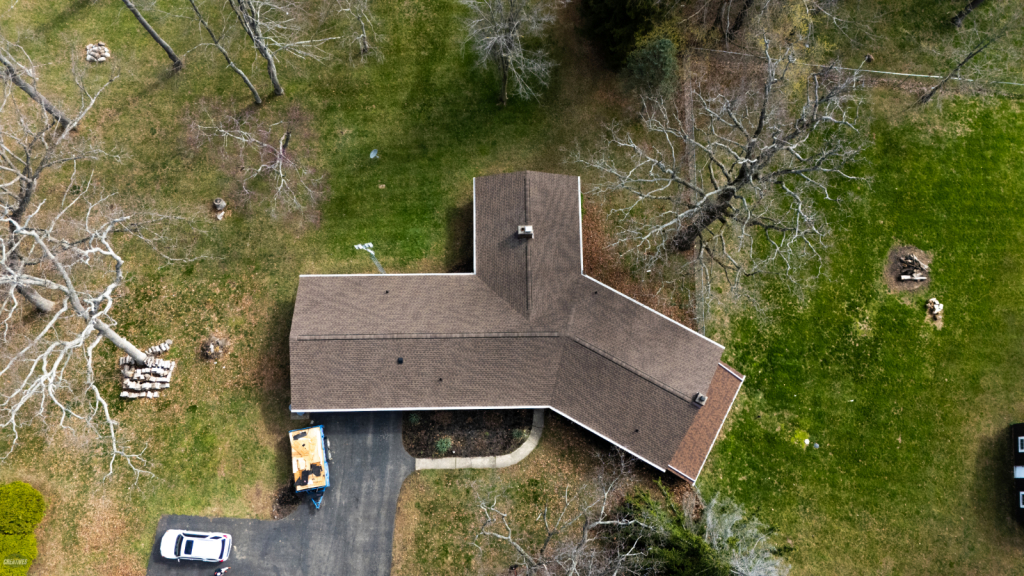 Aerial view of ranch-style house with brown shingle roof surrounded by green lawn and bare trees in yard