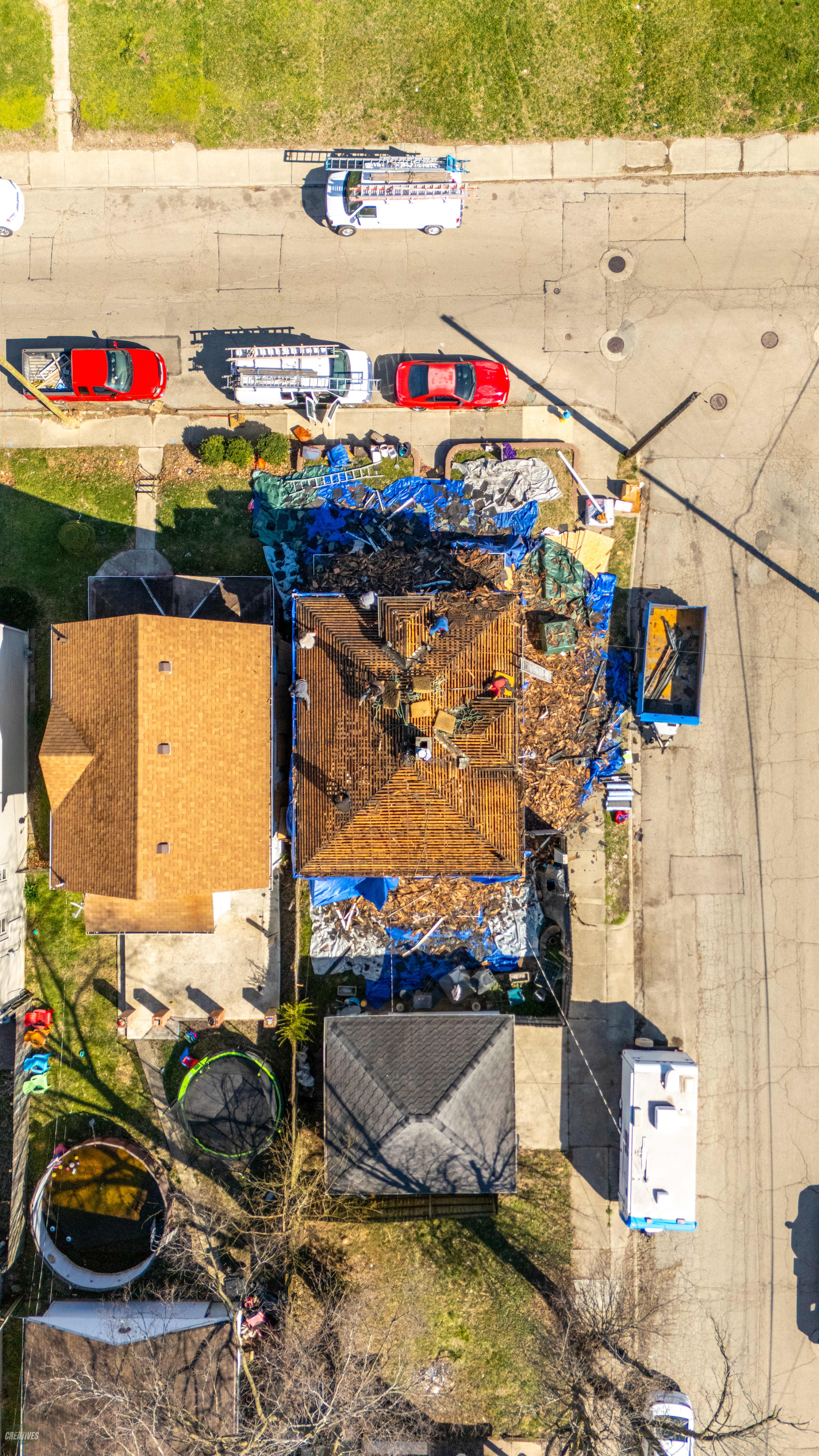 Aerial view of storm-damaged house with destroyed roof, blue tarps covering debris, emergency vehicles on street