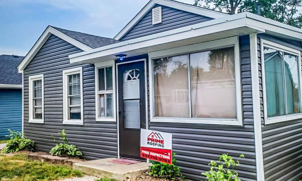 Gray vinyl siding house with white trim, large front window, and red Elite Roofing yard sign in front yard