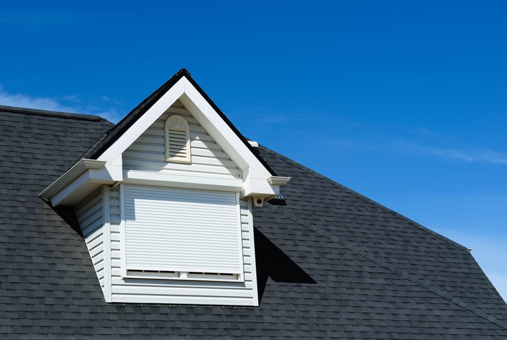 White dormer window with shutters on gray asphalt shingle roof against bright blue sky