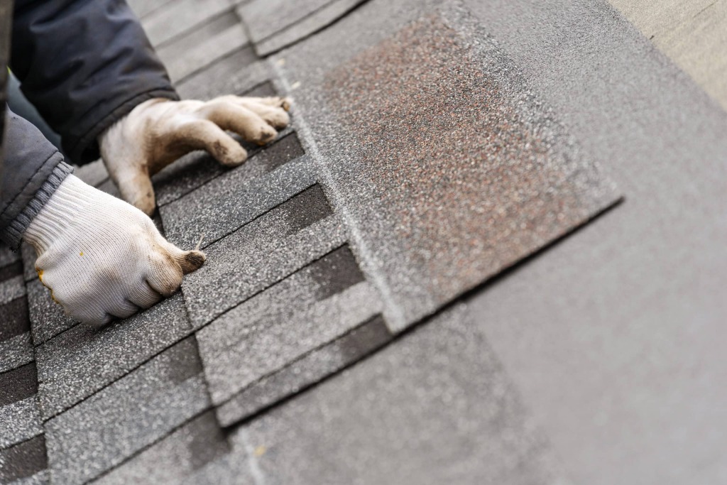 Professional roofer installing asphalt shingles on residential roof, hands positioning gray roofing materials