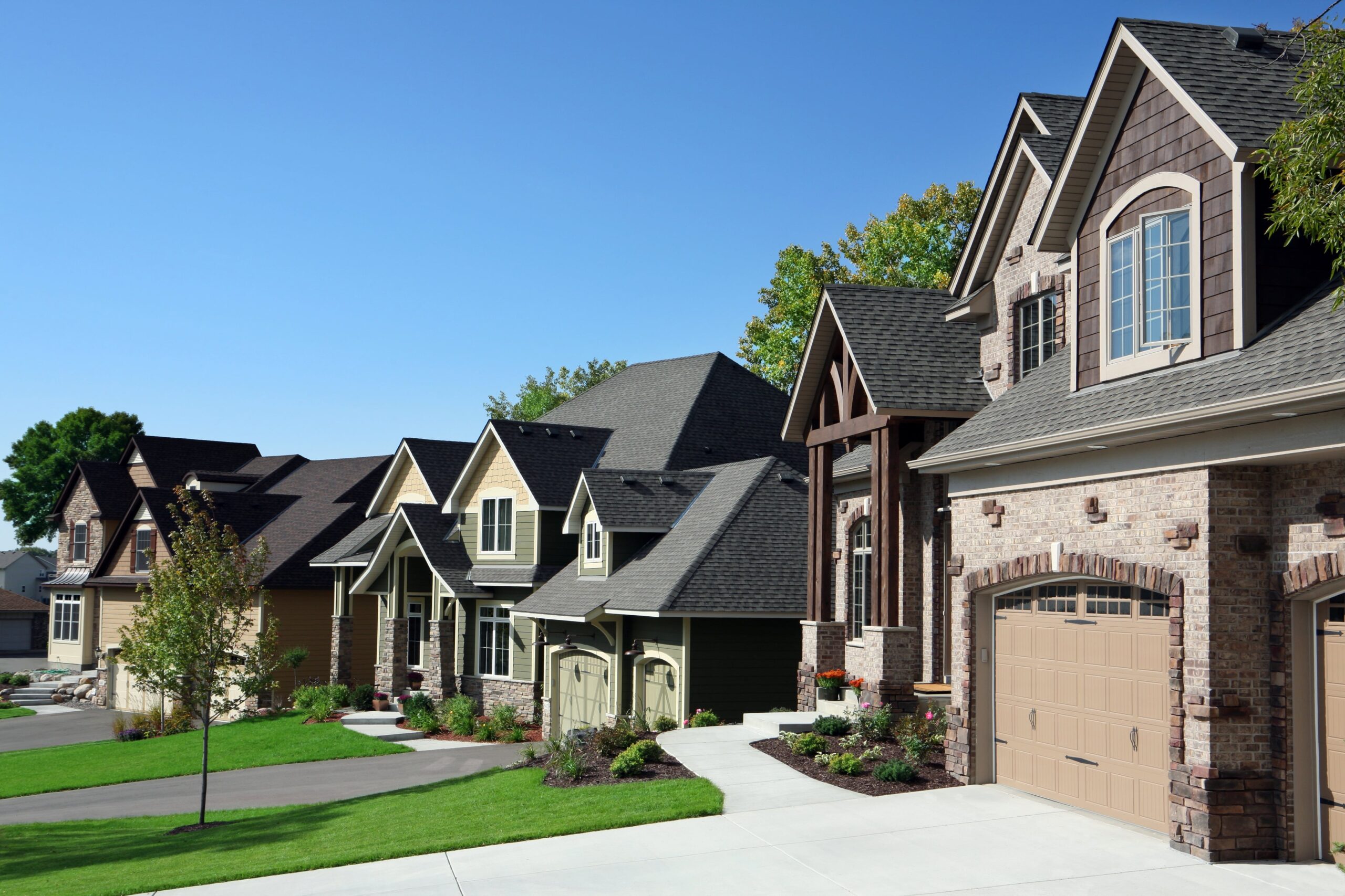 Luxury suburban homes with brick and stone exteriors, manicured lawns, and attached garages under clear blue sky