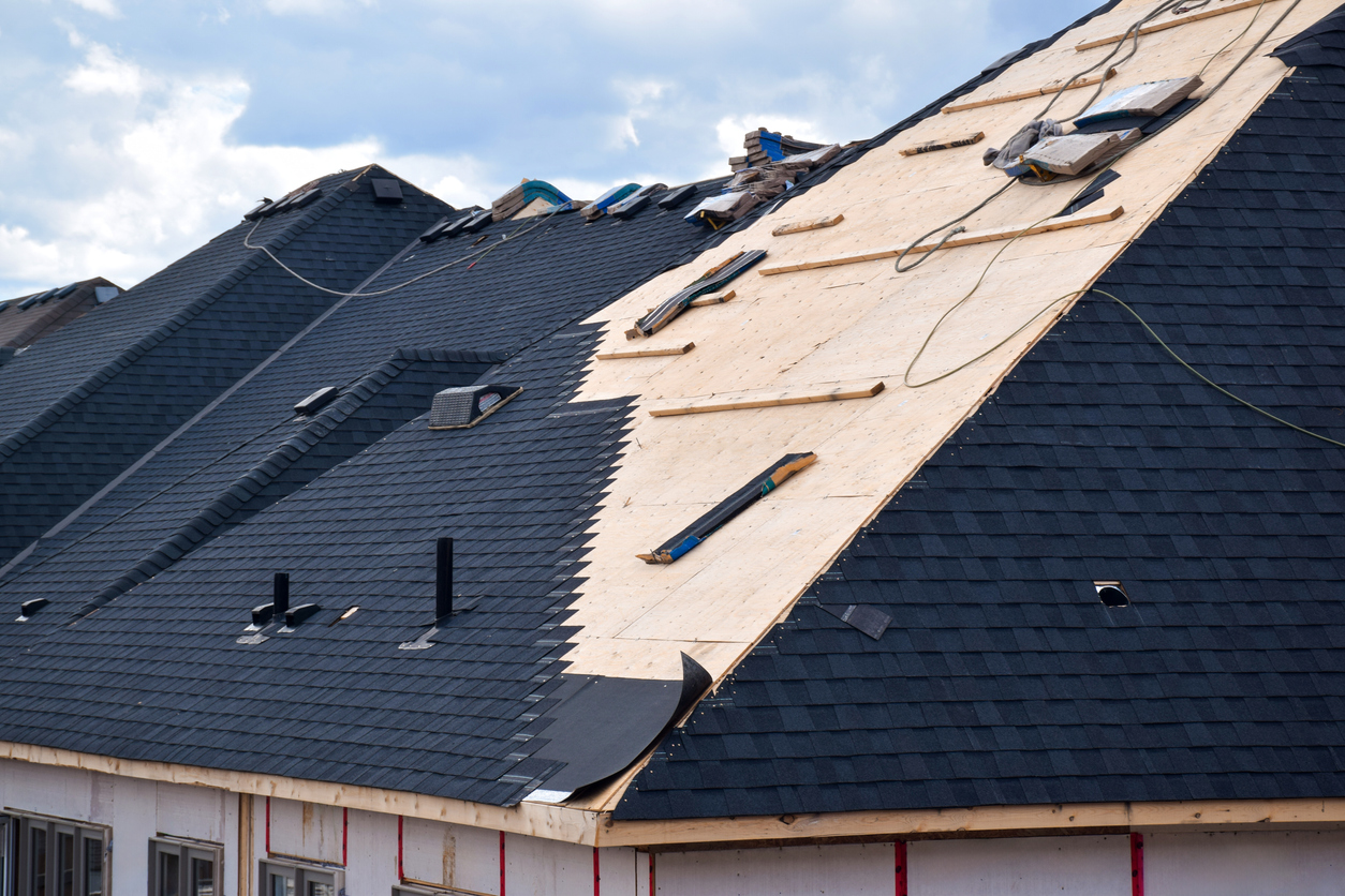 House roof under construction showing dark shingles and exposed plywood decking with roofing tools scattered