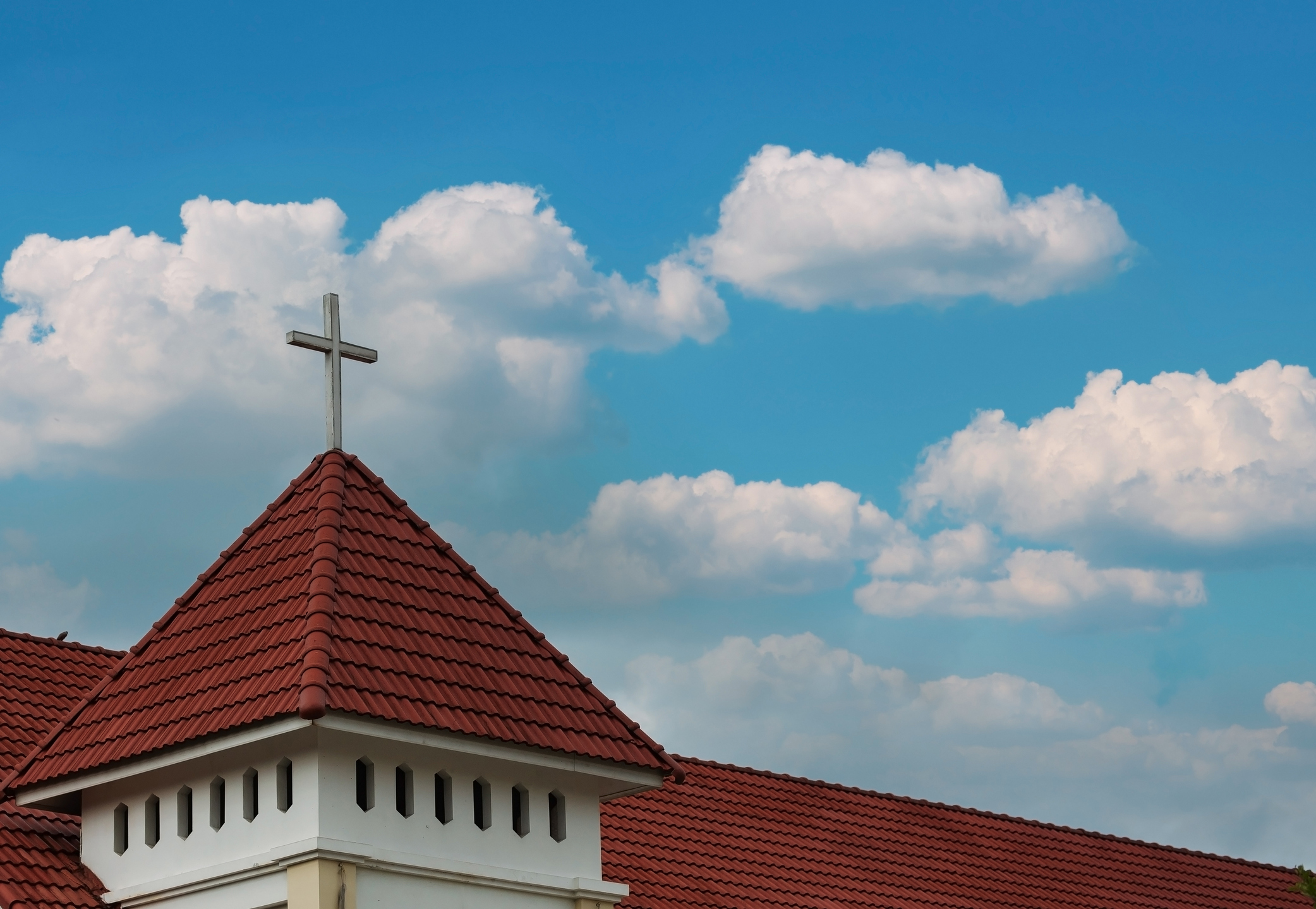 Church steeple with red tile roof and white cross against blue sky with fluffy white clouds