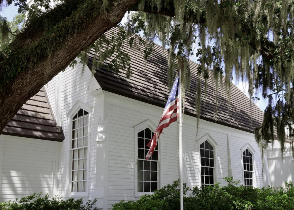 Historic white clapboard church with arched windows and wooden shingle roof under Spanish moss draped oak tree