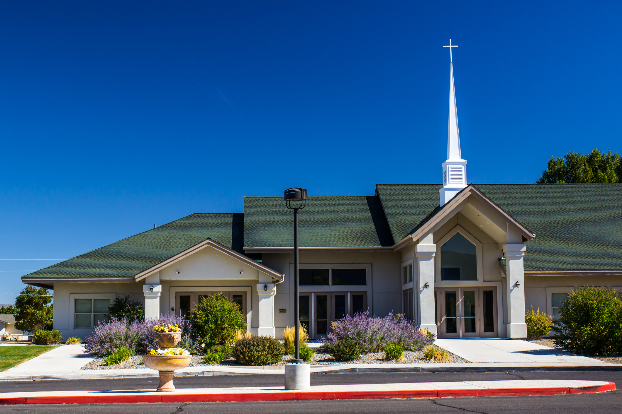 Modern church building with white steeple and cross, beige exterior, green roof, and landscaped front yard