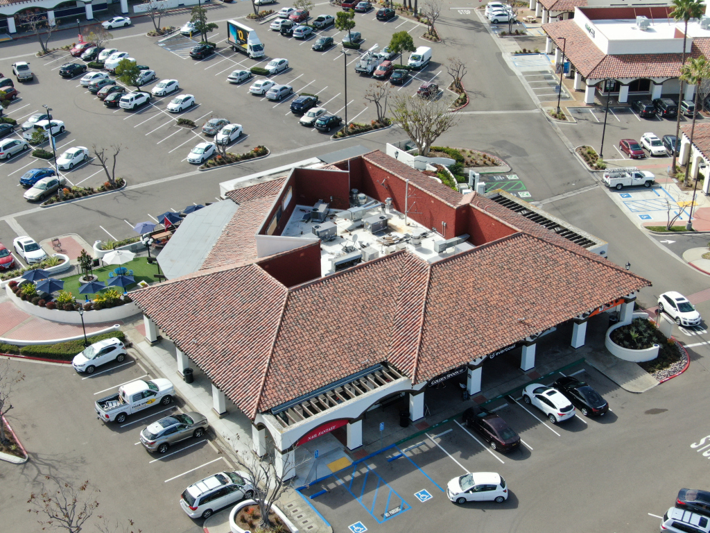 Aerial view of shopping center with red tile roof building, large parking lot filled with cars, and landscaping