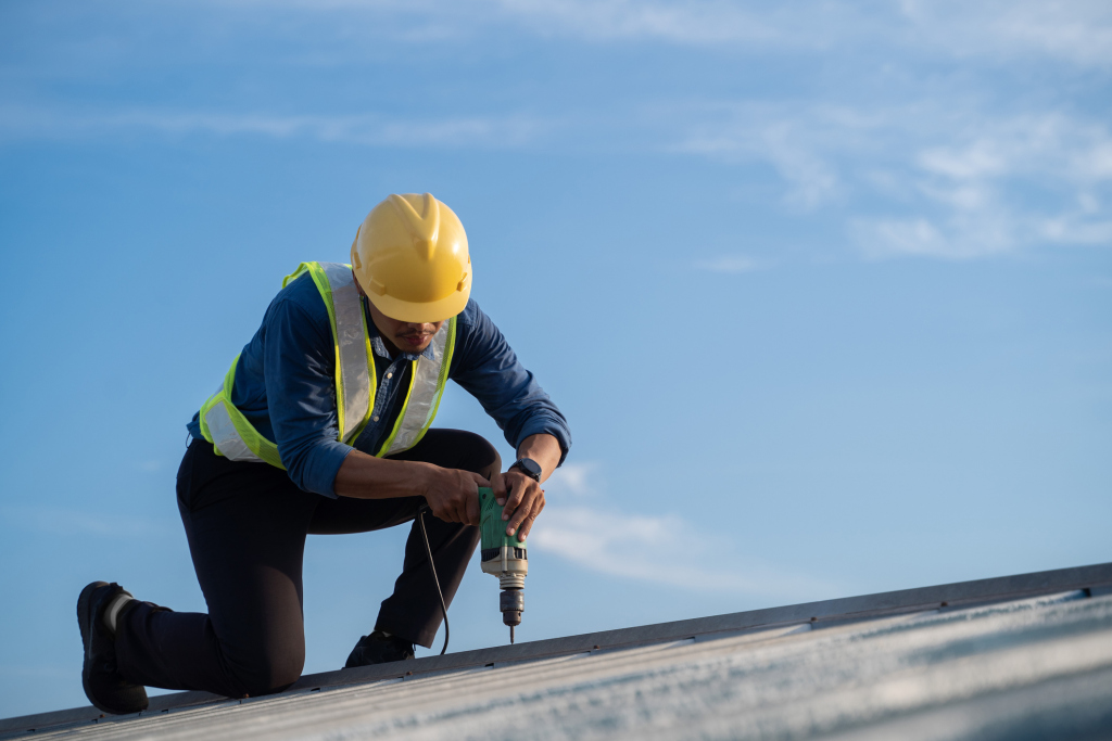 Construction worker in yellow hard hat and safety vest using power drill on roof against blue sky background