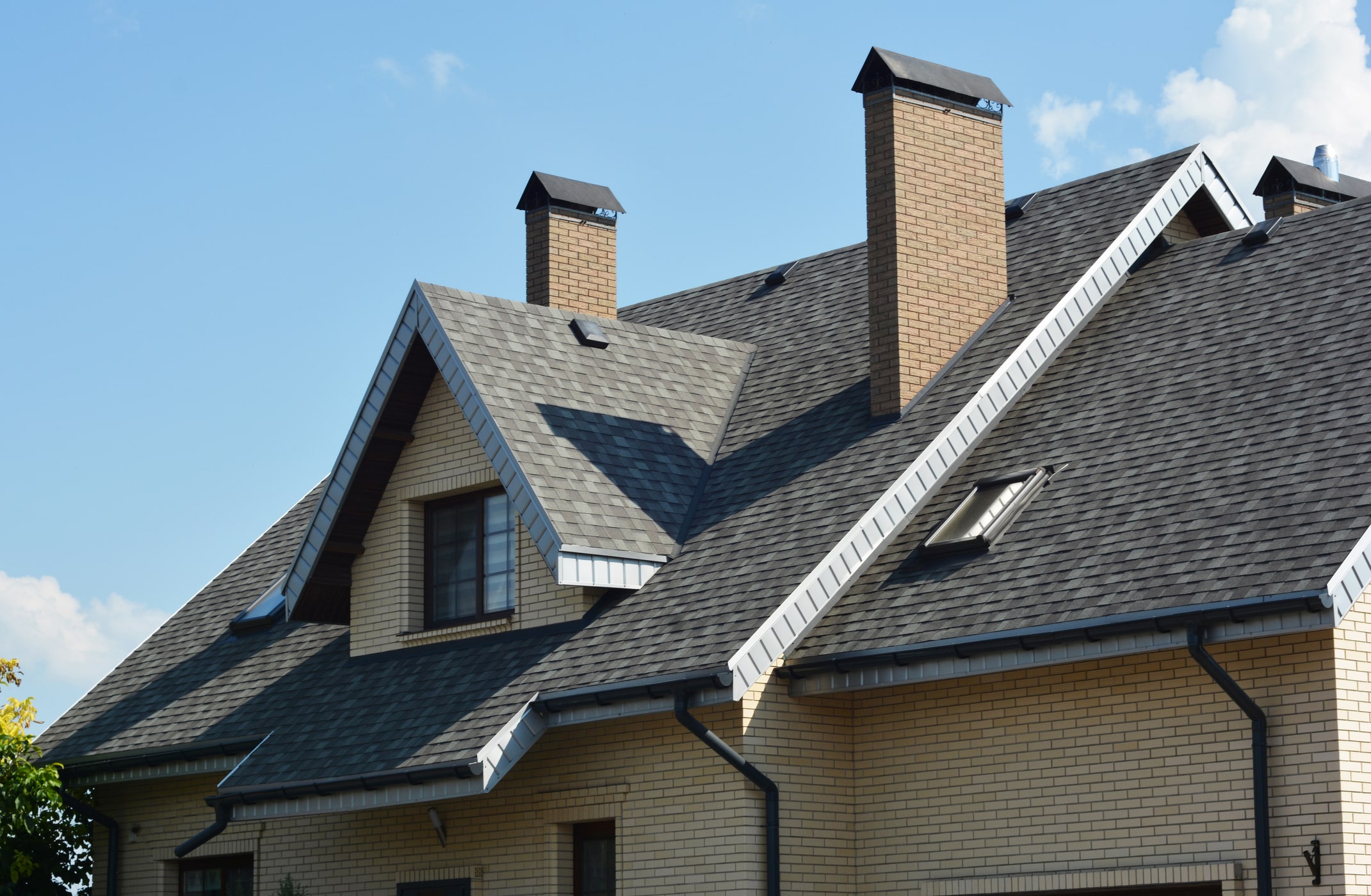 Modern brick house with dark gray shingle roof, multiple chimneys and dormer windows against blue sky