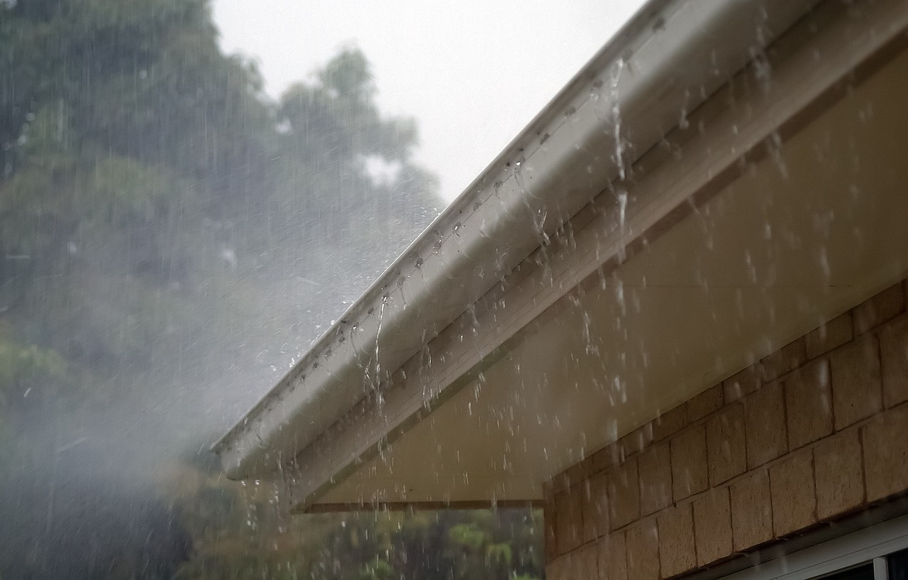 Heavy rain pouring from roof gutter on residential house with shingle siding during storm with trees in background