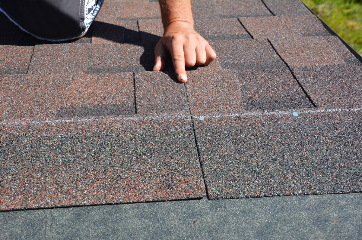 Hand pointing to damaged asphalt roof shingles showing cracks and wear during professional roof inspection