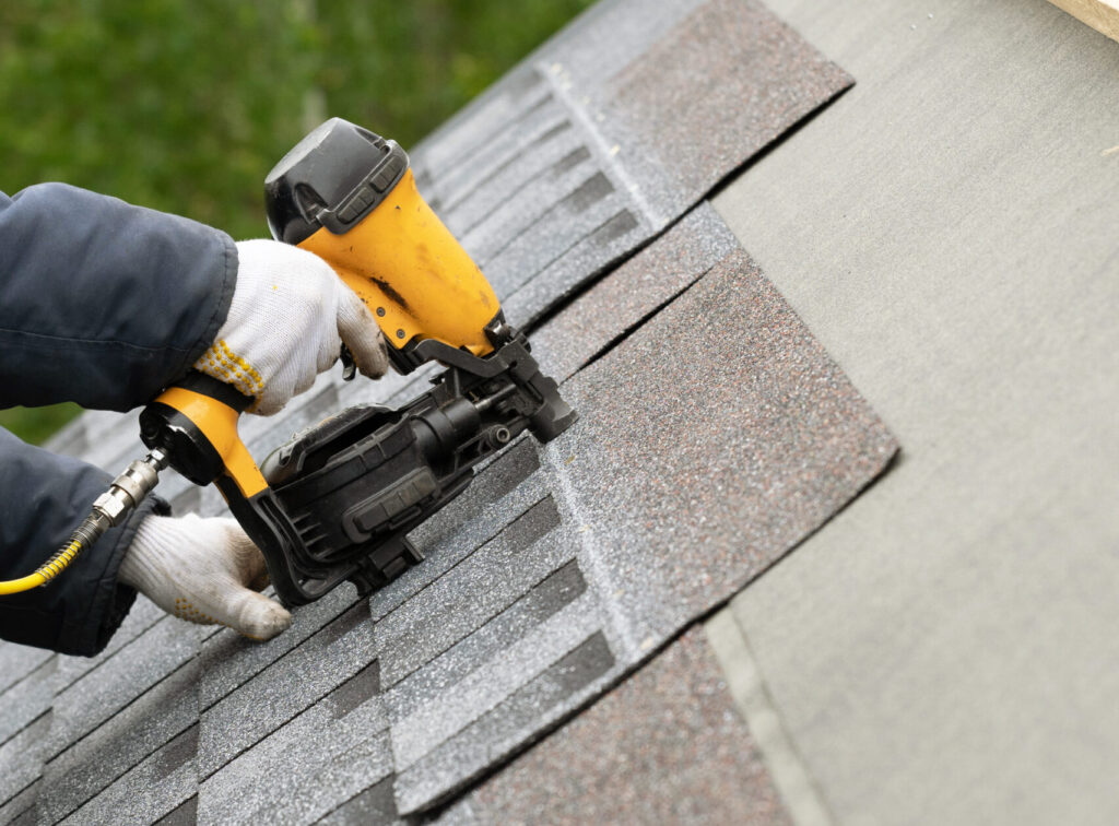 Roofer using yellow pneumatic nail gun to install asphalt shingles on residential roof with protective gloves