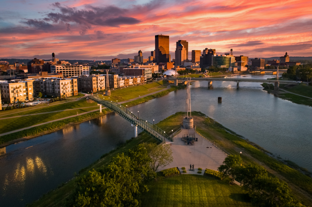 Aerial view of downtown city skyline at sunset with river, pedestrian bridge, and waterfront park in golden light