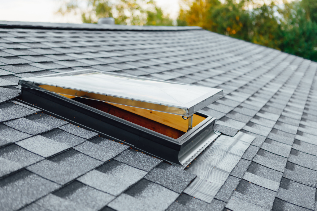 Open roof skylight with metal flashing installed on gray asphalt shingle roof with autumn trees in background