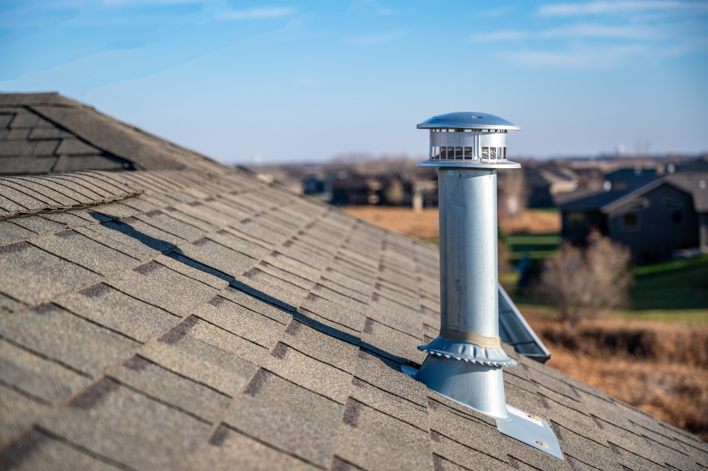Silver metal chimney pipe with cap installed on brown asphalt shingle roof with residential neighborhood background