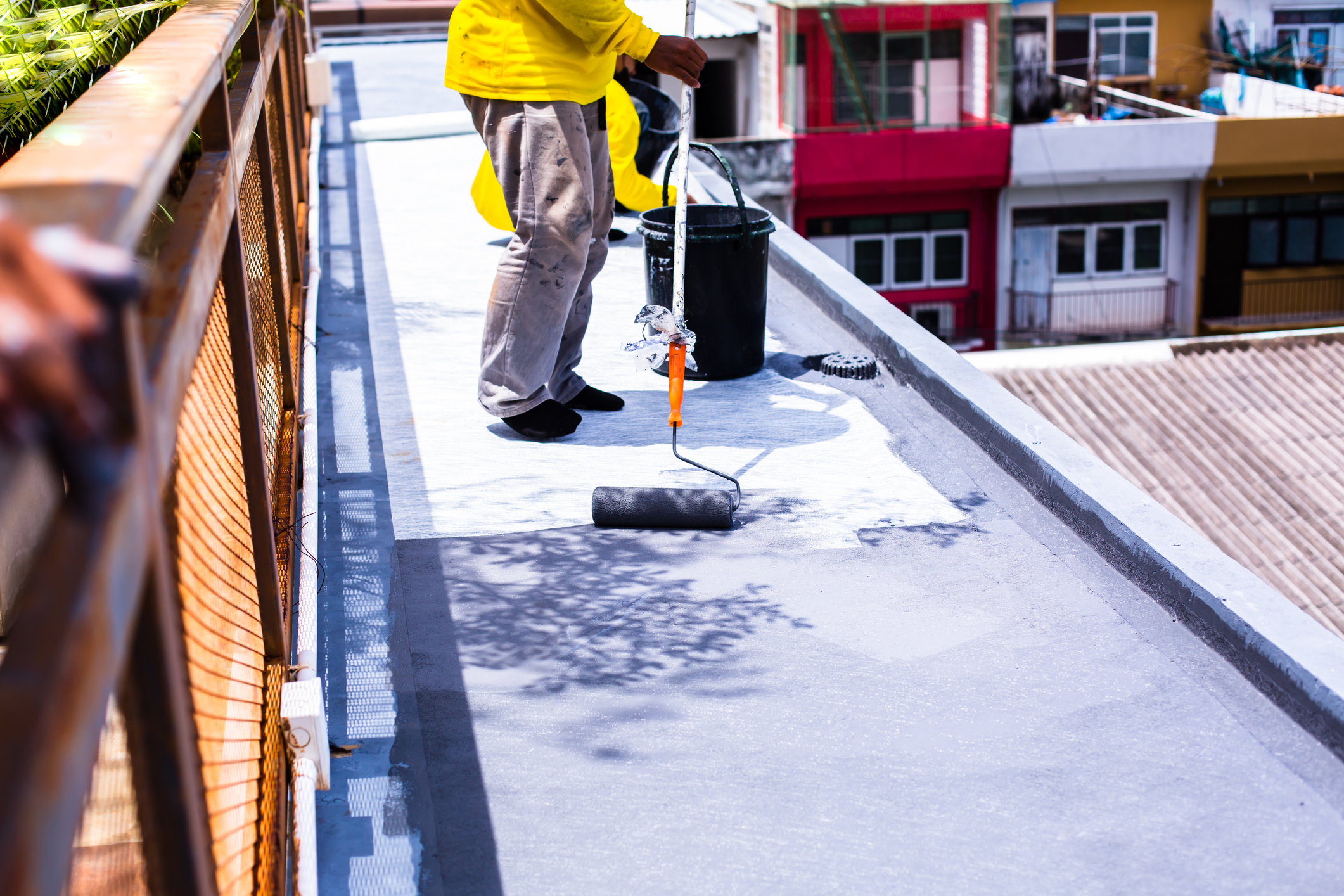 Worker in yellow shirt applying waterproof coating to rooftop with paint roller and bucket on residential building