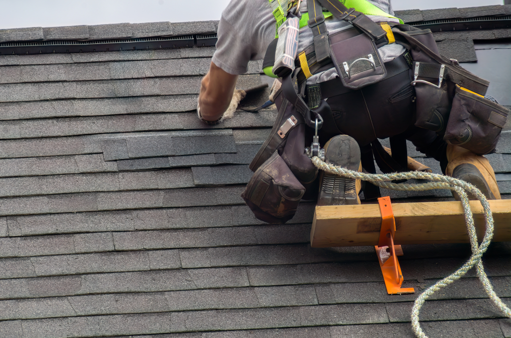 Roofer in safety harness installing shingles on residential roof with tool belt and safety equipment