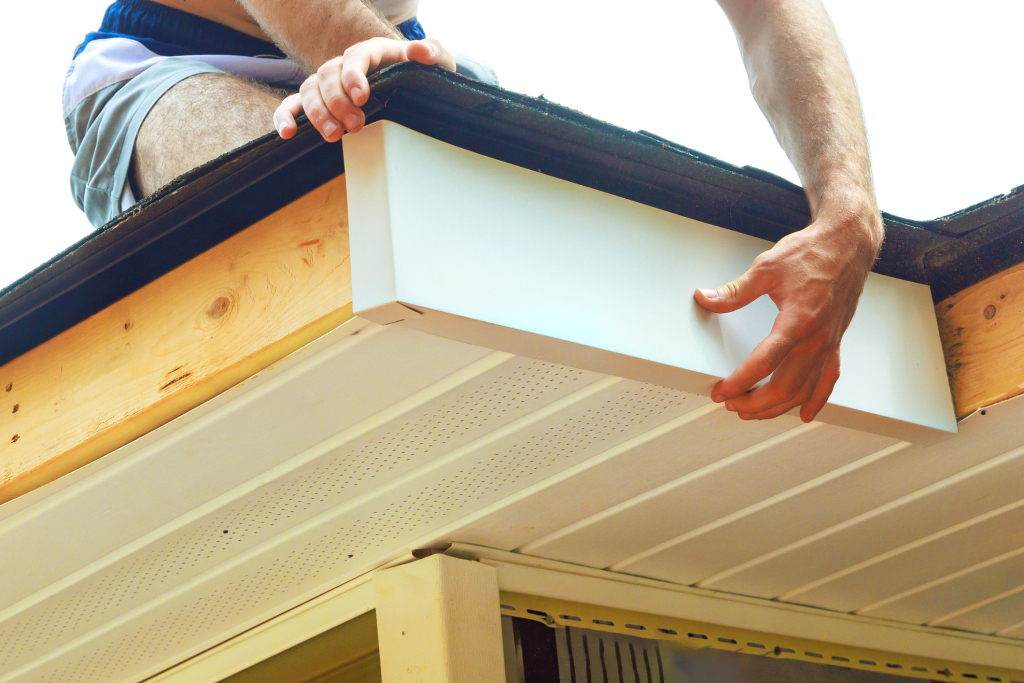 Worker installing white vinyl soffit panels on house eaves with wooden rafters visible above