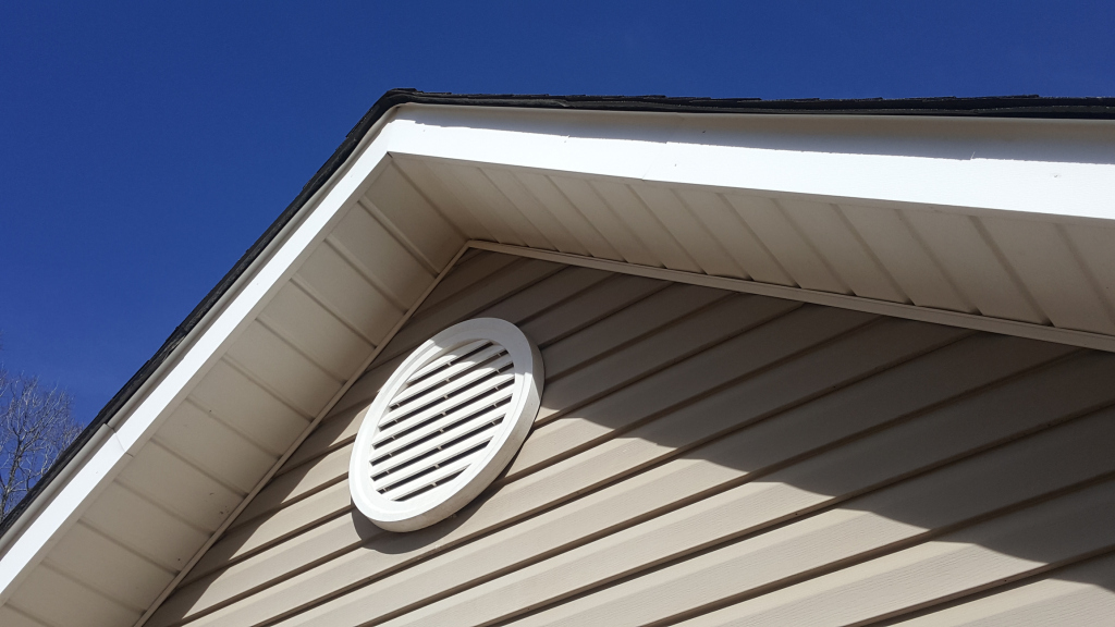 Close-up view of house gable with circular white ventilation vent on beige vinyl siding under clear blue sky