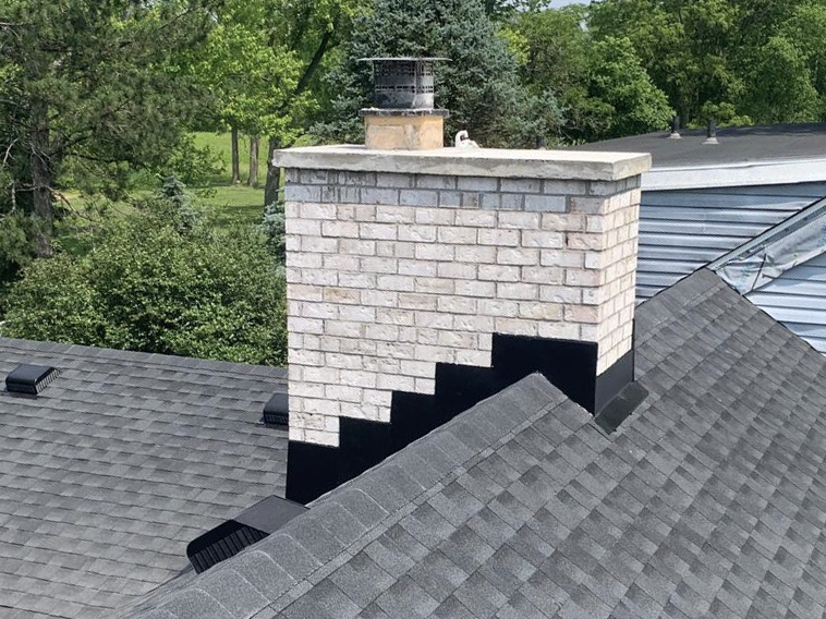 White brick chimney with metal cap on gray shingle roof surrounded by lush green trees in residential area