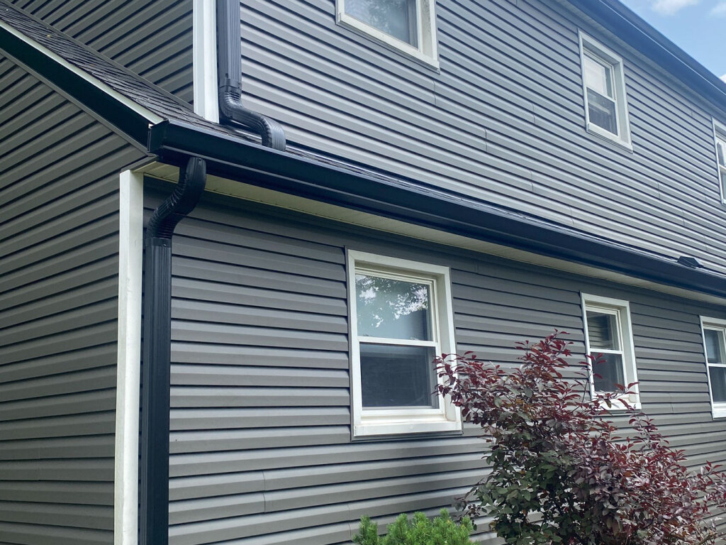 Gray vinyl siding house exterior with dark gutters and downspouts, white-trimmed windows, and landscaping