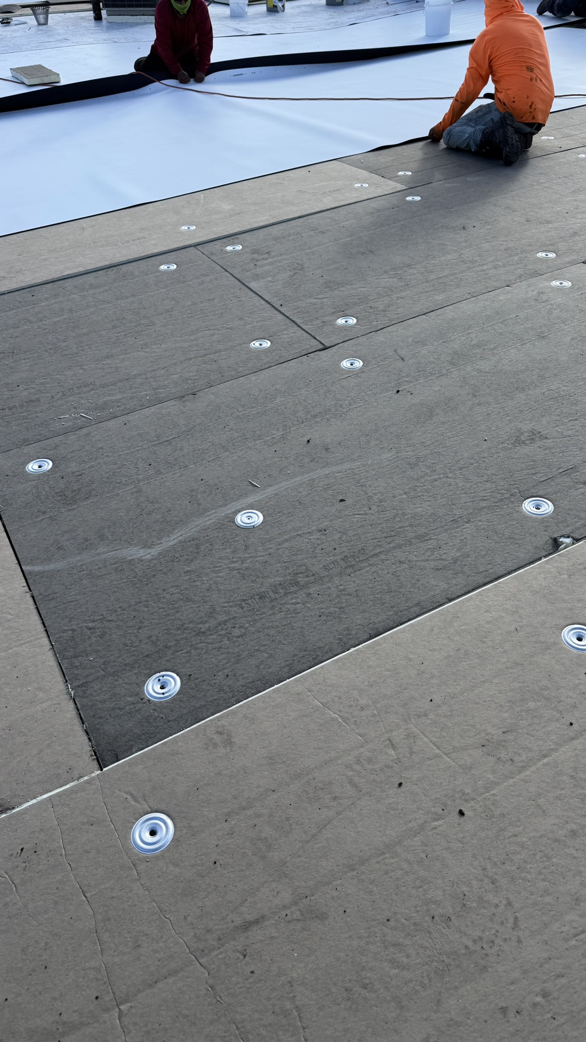 Construction workers installing white membrane roofing material on concrete deck with circular fasteners