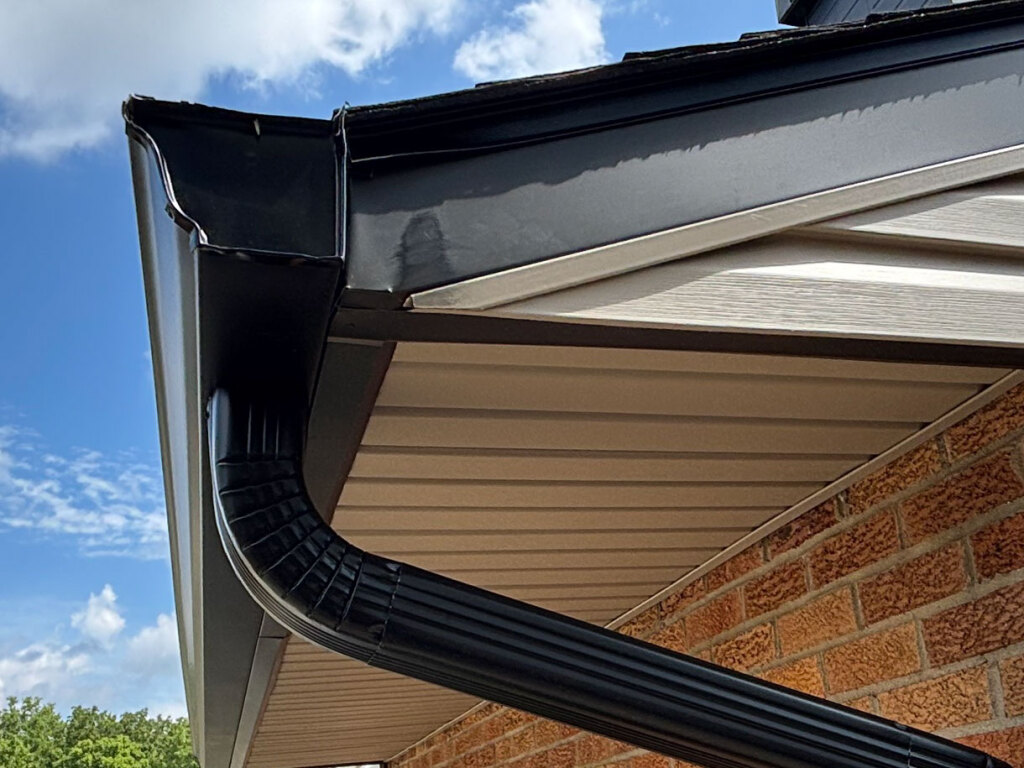Black seamless gutters and downspout system installed on brick house with beige vinyl siding under blue sky