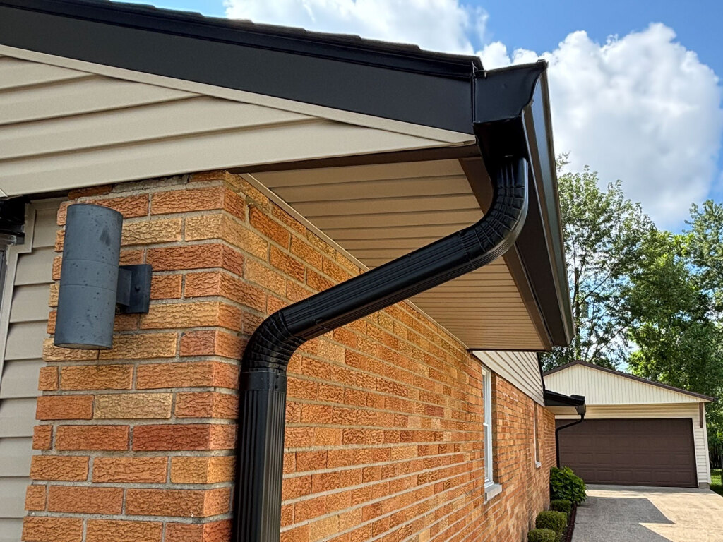 Black rain gutter system installed on brick house exterior with beige siding and garage in background