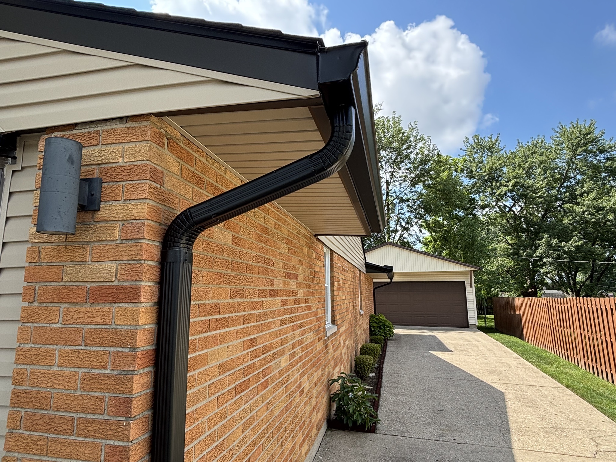 Modern brick house exterior with dark gutters, concrete driveway, and wooden fence under blue sky with clouds