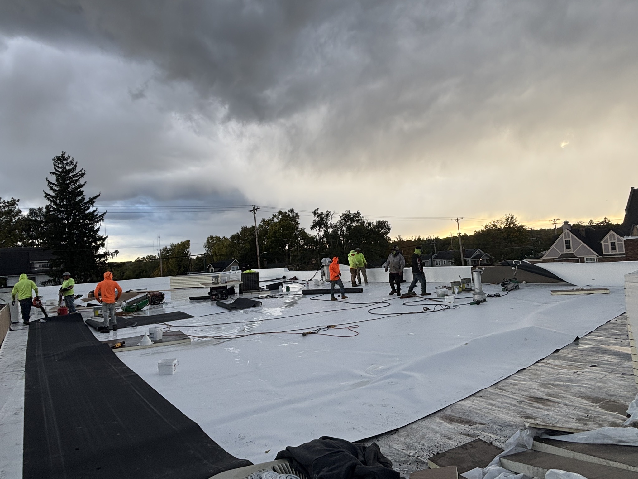 Construction workers in hi-vis safety gear installing white roofing membrane on commercial building under stormy sky