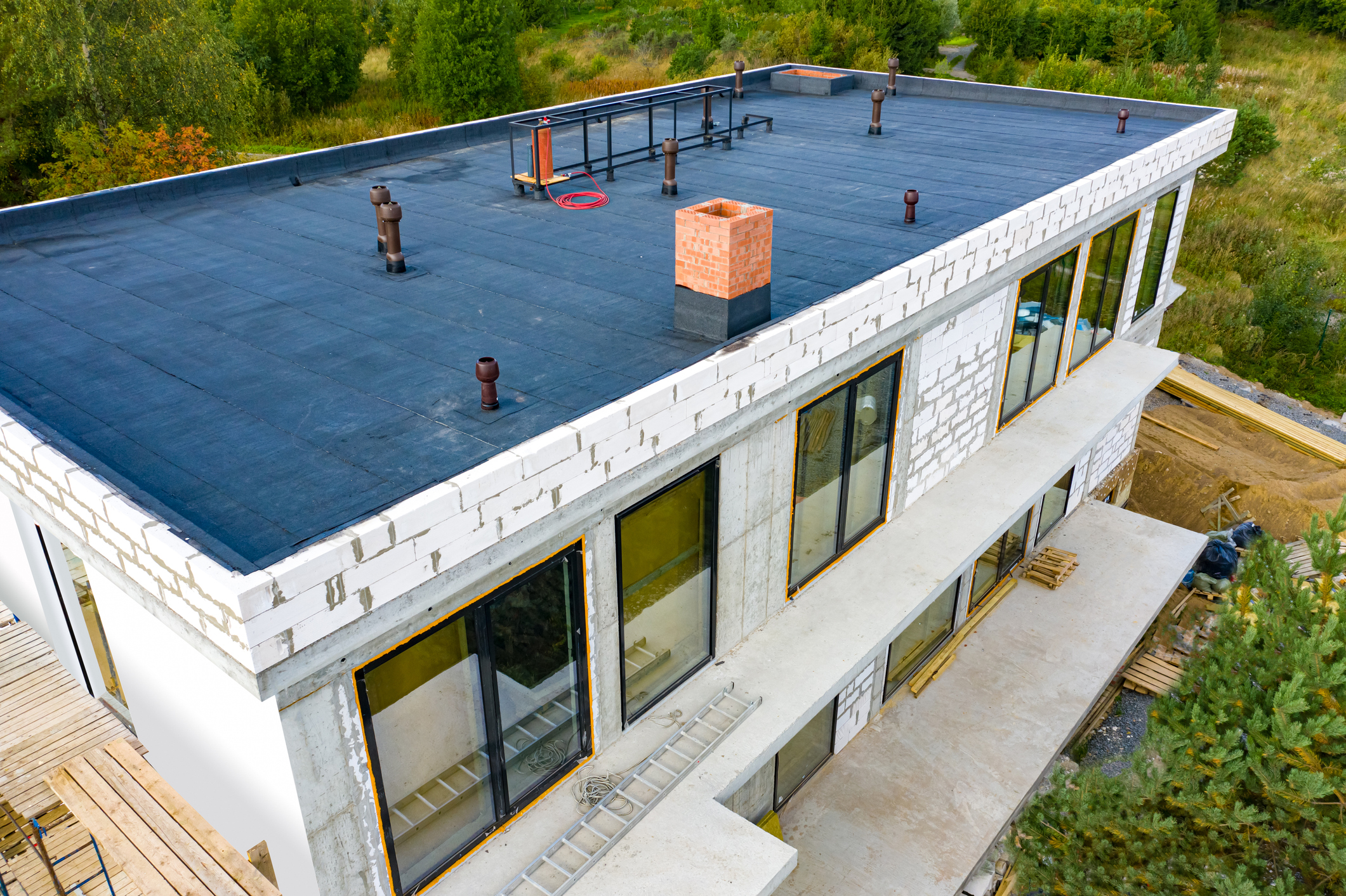 Aerial view of modern house under construction with flat roof, brick chimney, and large windows surrounded by forest