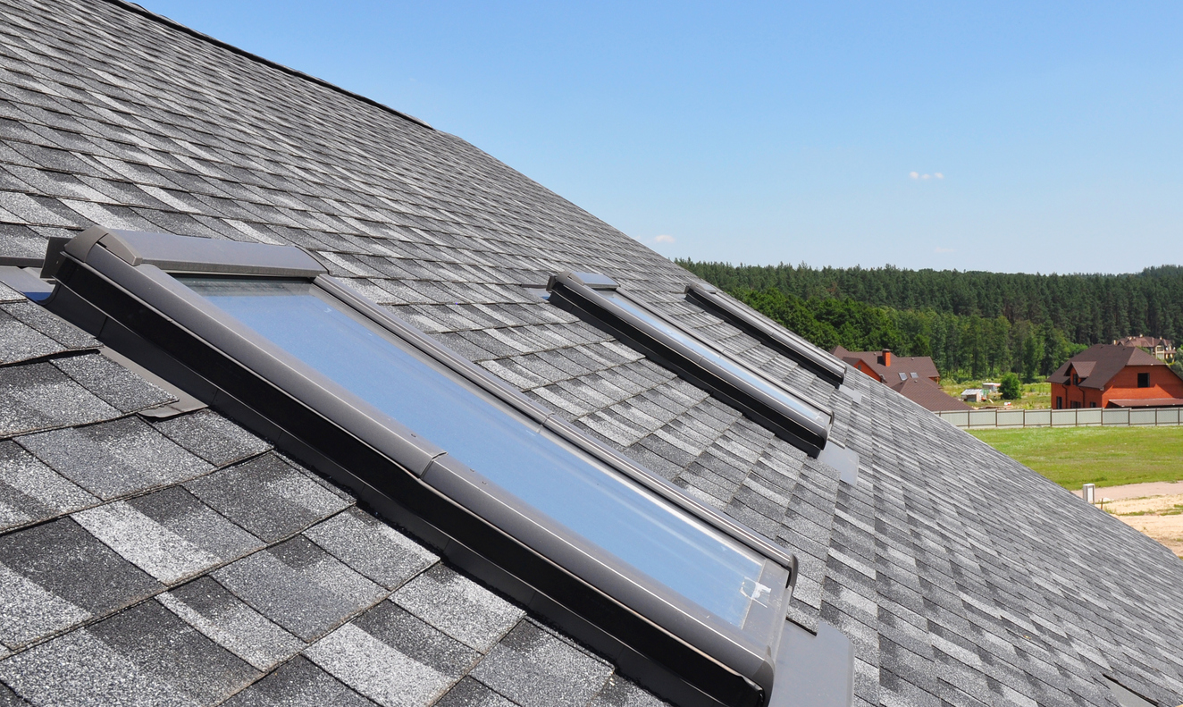 Modern skylight windows installed on gray shingle roof with forest and residential homes in background