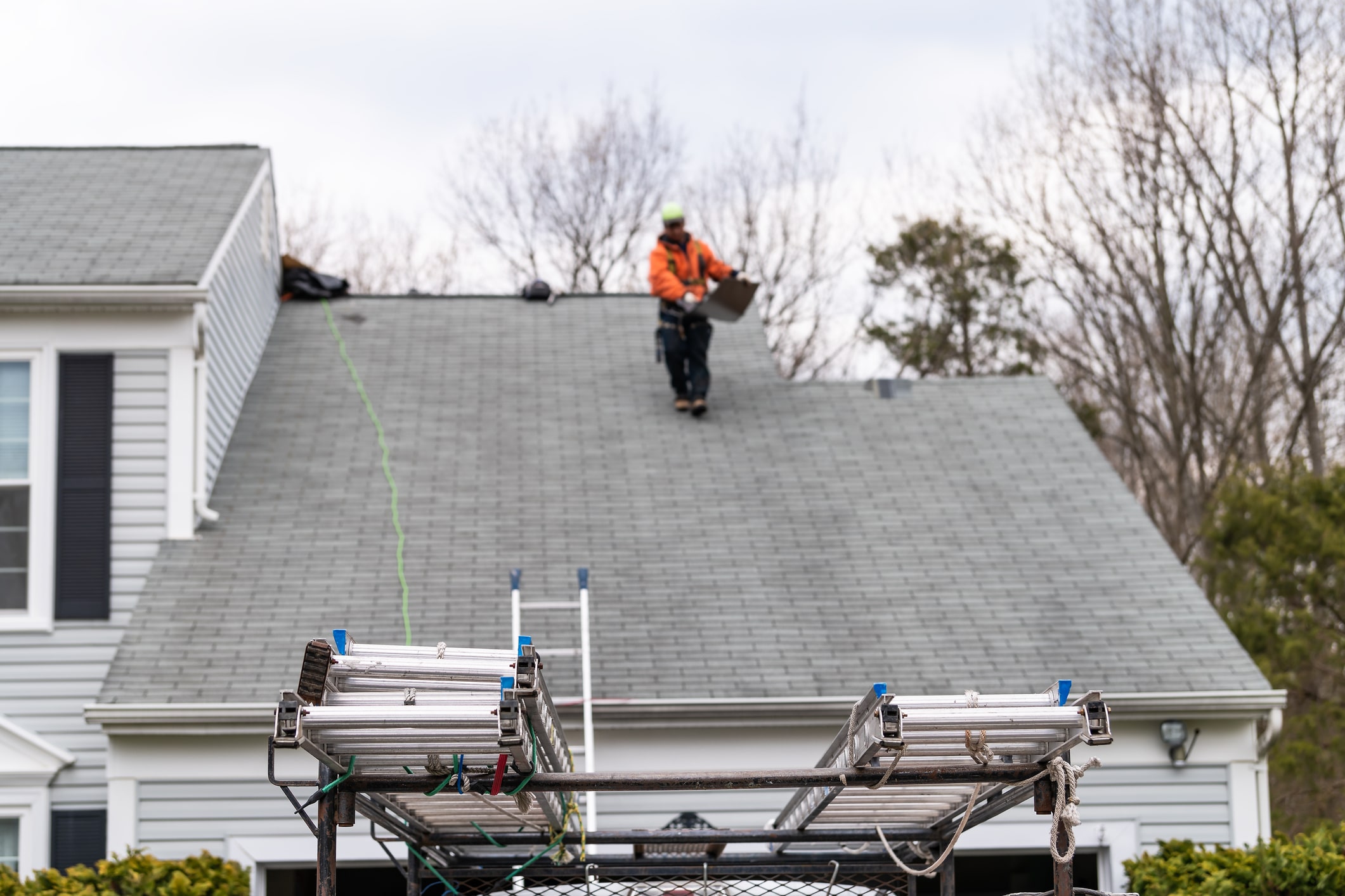 Roofer in orange safety vest working on gray shingle roof with aluminum scaffolding below and white house