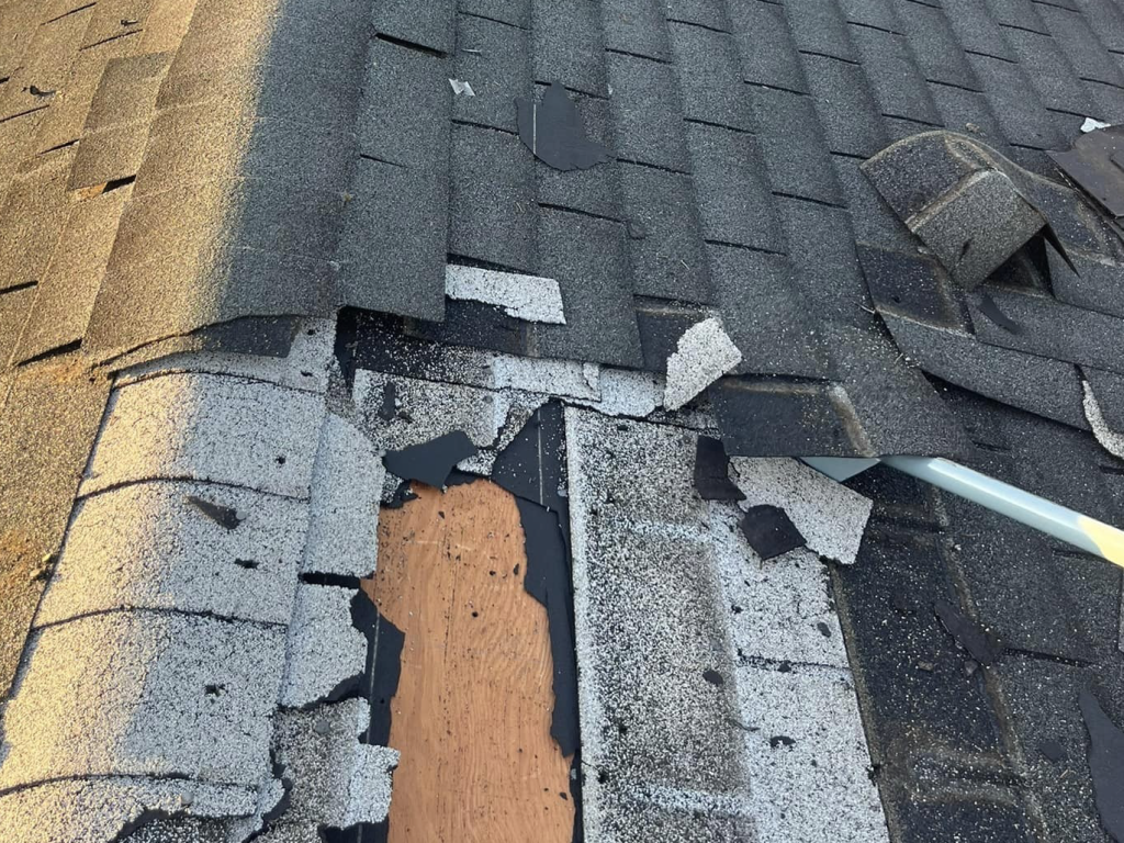 Damaged asphalt shingles on roof with exposed wood underlayment and debris, showing storm or weather damage
