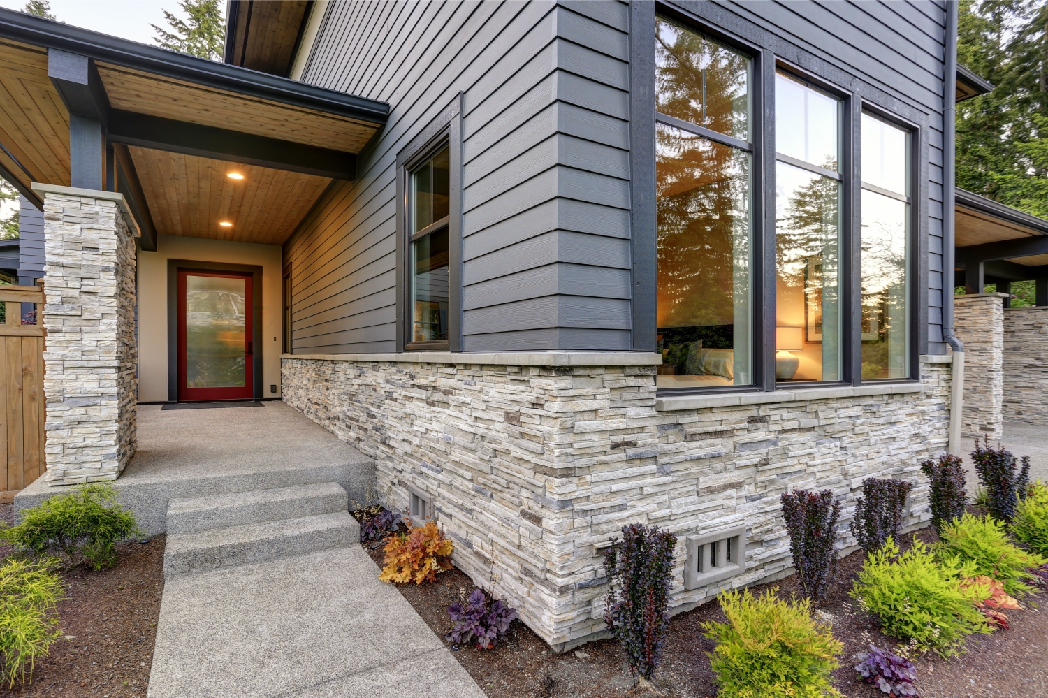 Modern home exterior with gray siding, stacked stone foundation, large windows, and landscaped entrance