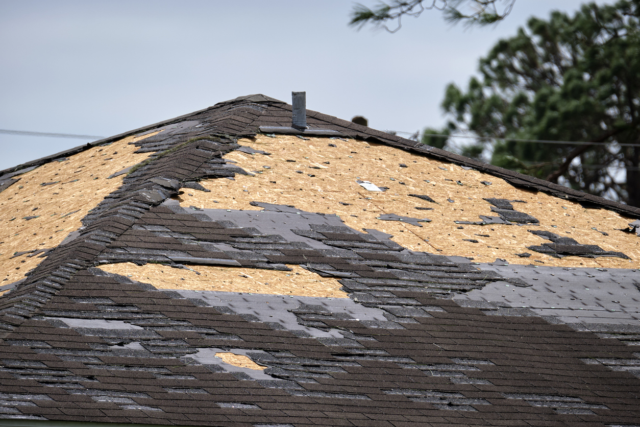 Damaged roof with missing shingles exposing plywood underlayment, showing severe storm or weather damage repair needs